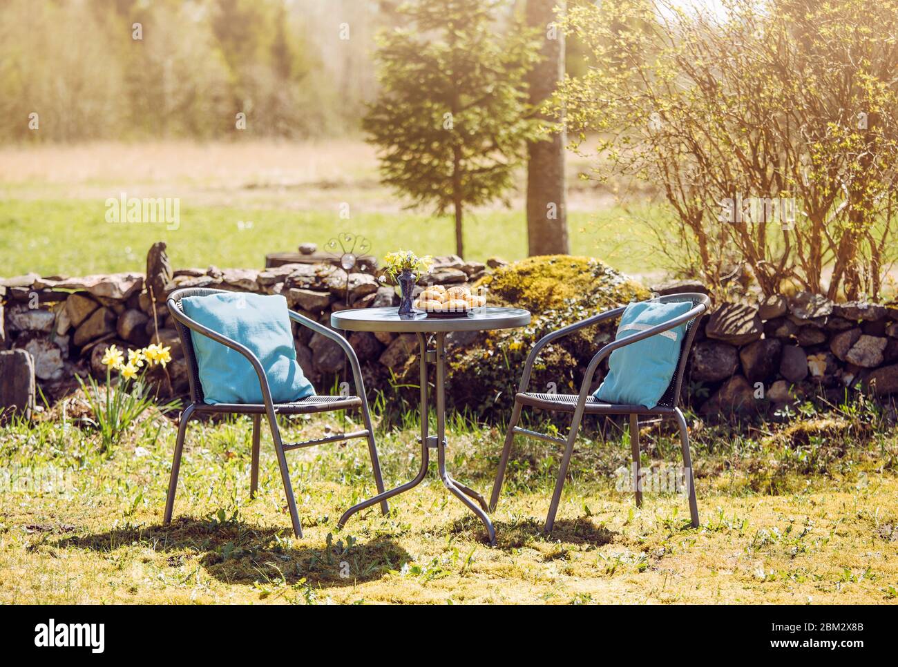 Kleiner Tisch im ländlichen Garten mit Stühlen und Tisch für zwei, frisch gebackenes Gebäck auf dem Tisch, sonniger Frühlingsmorgen. Gestapelte Steinzaun Wand an Stockfoto