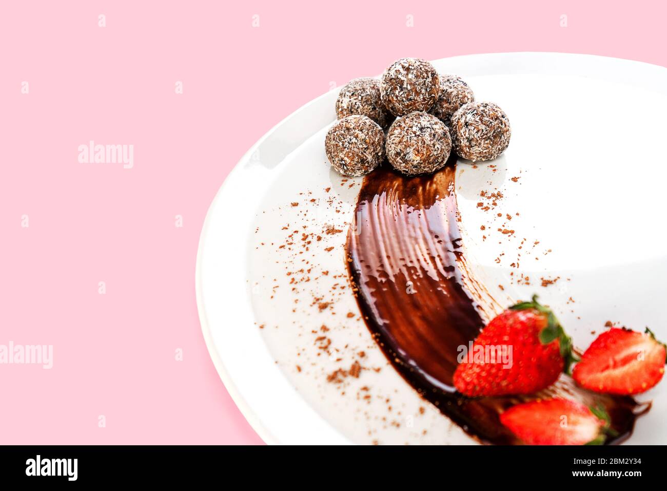 Trendige Energiebälle oder Energiebisse mit Hafer, Schokolade und Kokosnuss auf weißem Teller mit Erdbeere. Gesunde vegane und rohe Ernährung. Selektiver Fokus. Kopierbereich. Stockfoto