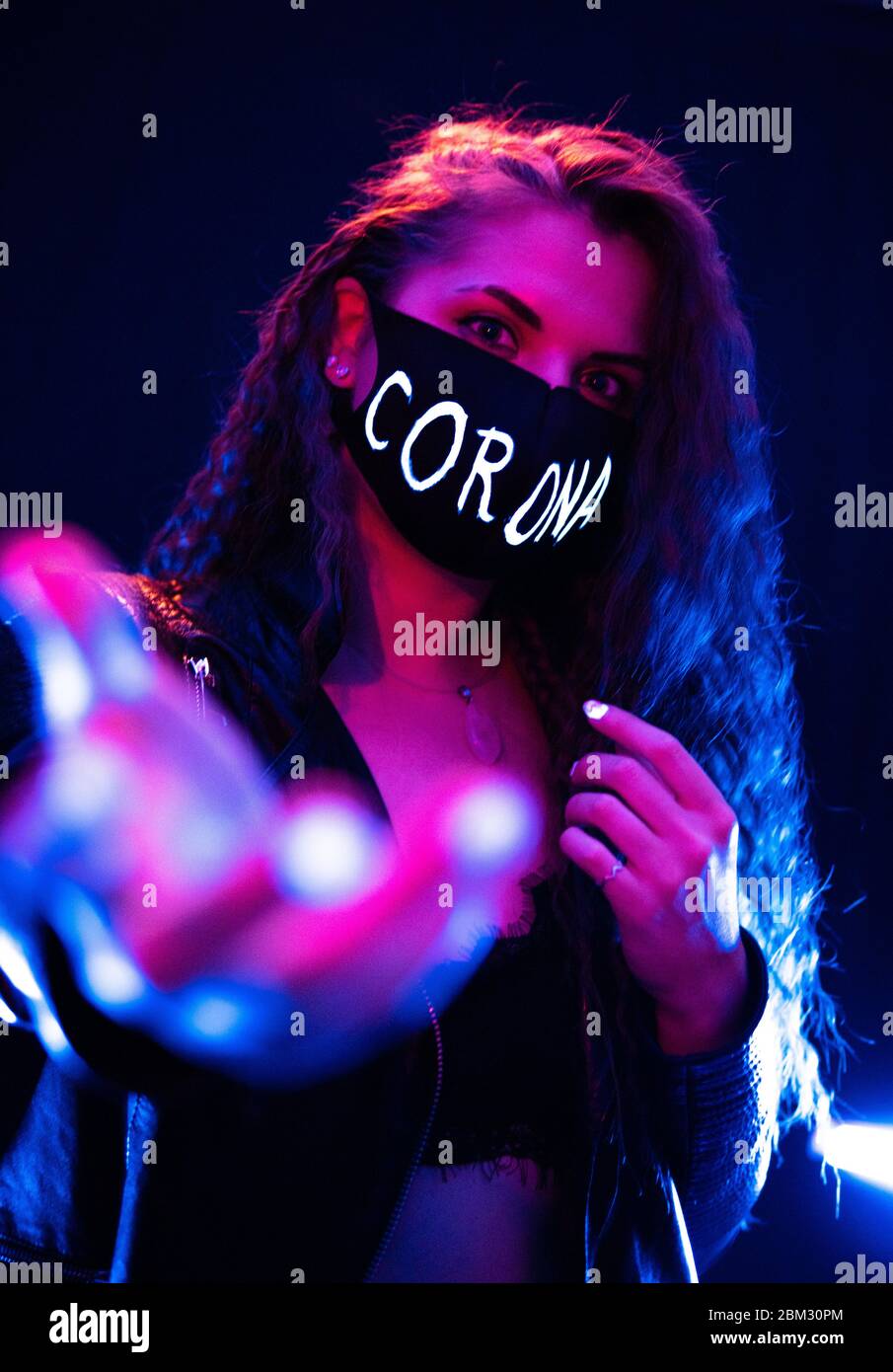 Porträt eines winkenden Attraktive Mädchen trägt schwarze Schutzmaske mit Corona Zeichen auf Gesicht in Neon Lights mit schwarzem Foggy Hintergrund Stockfoto