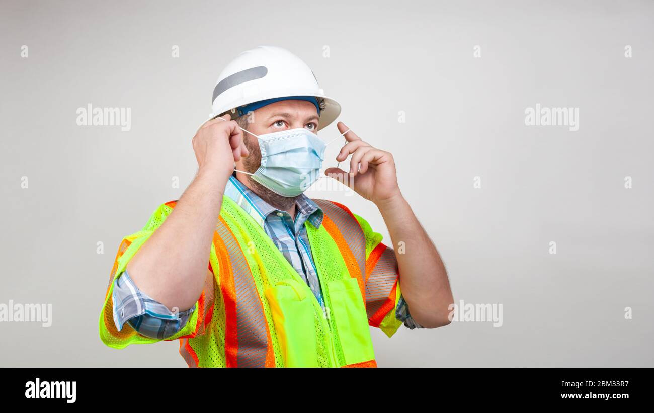 Bauarbeiter mit Schutzhelm und Sicherheitsweste setzt schützende Operationsmaske auf. Konzept der Maßnahmen zur Verlangsamung der Ausbreitung von Covid-19 Stockfoto