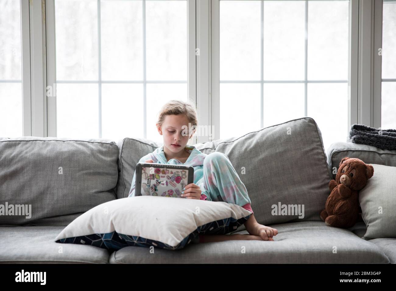 Das blonde Mädchen aus der Schule spielt Tablette auf der Couch mit Teddy Stockfoto
