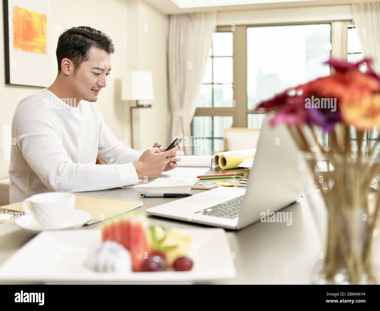 Junge asiatische Mann Design professionelle Arbeit von zu Hause aus sitzen in der Küche mit Handy (Kunstwerk im Hintergrund digital verändert) Stockfoto