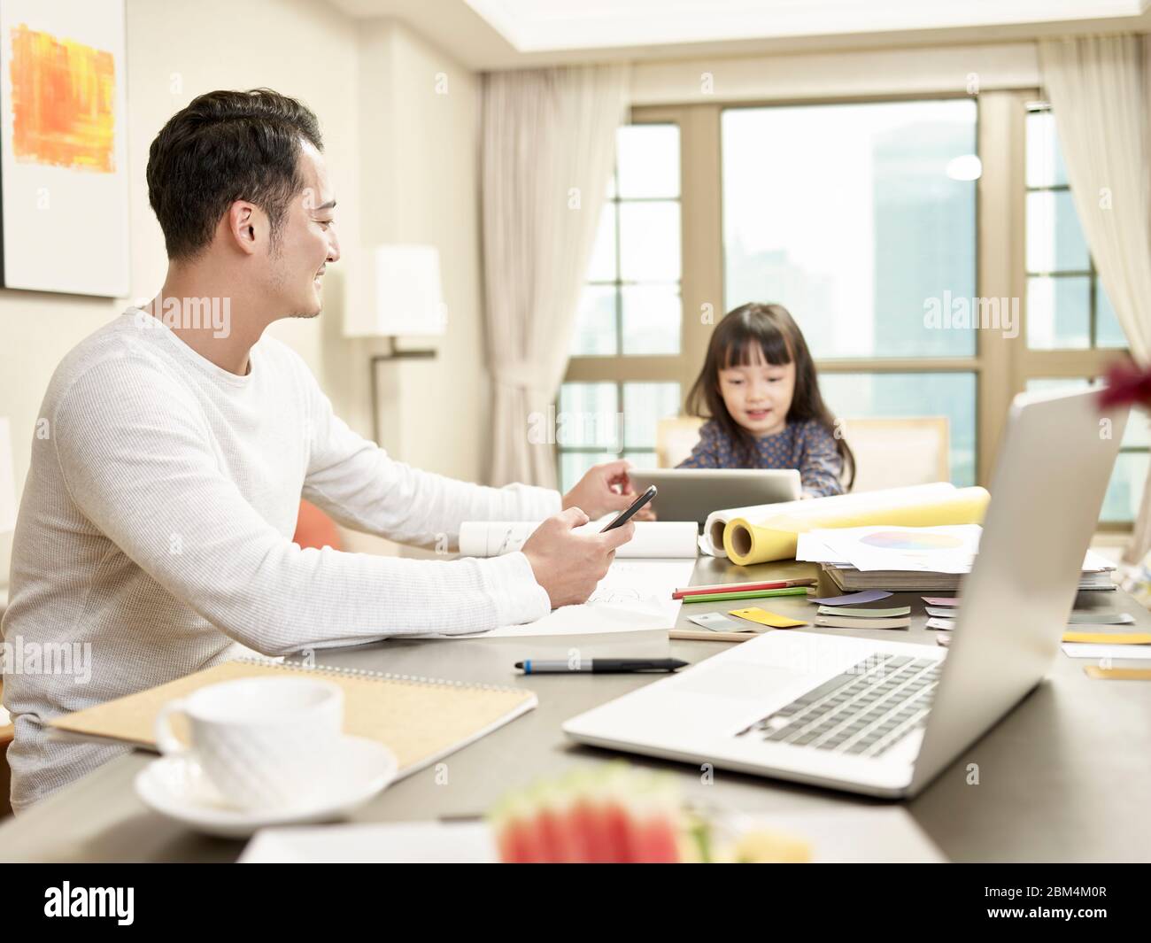 Junge asiatische Geschäftsmann kümmert sich um Kind während der Arbeit von zu Hause aus (Kunstwerk im Hintergrund digital verändert) Stockfoto