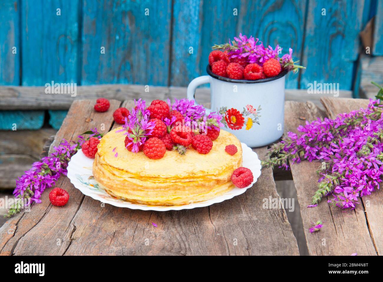 Stapel von frisch gebackenen Crepes mit Handvoll frischen Himbeeren auf der Oberseite. Alte Utensilien und Feldblumen auf Holztisch. Stockfoto