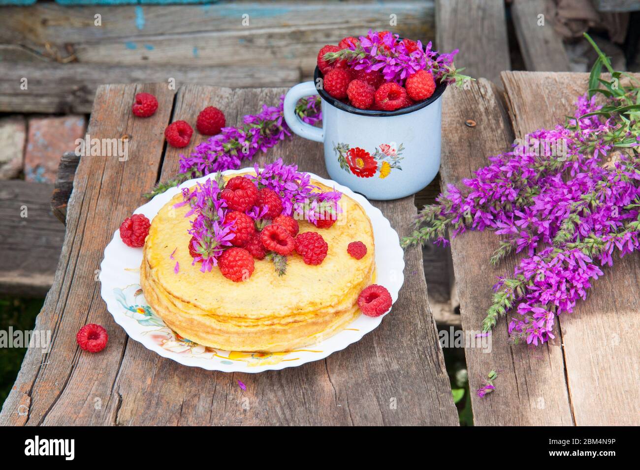 Stapel von frisch gebackenen Crepes mit Handvoll frischen Himbeeren auf der Oberseite. Alte Utensilien und Feldblumen auf Holztisch. Stockfoto