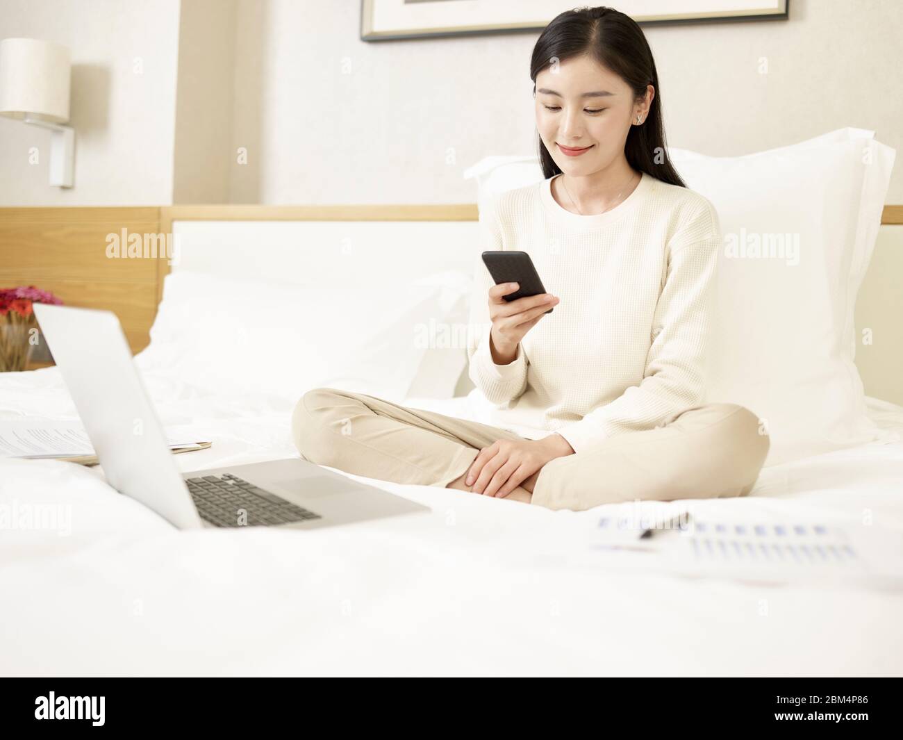 Schöne junge asiatische Frau sitzt auf dem Bett arbeiten mit Handy und Laptop Stockfoto