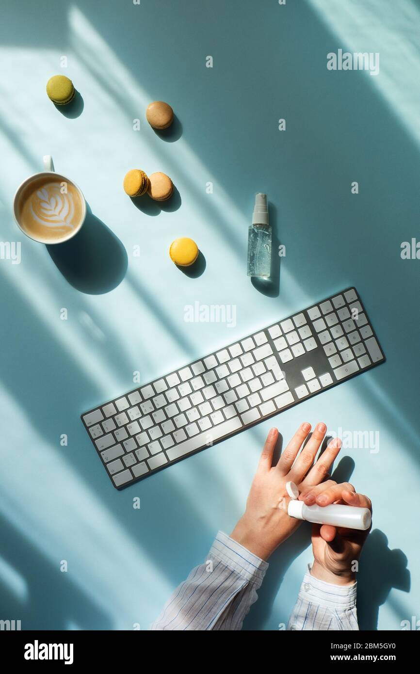 Arbeitsplatz zu Hause am Fenster. Flacher Lat mit einer Tasse Kaffee und Makronen im Sonnenlicht und Frauenhände mit Desinfektionsmittel. Stockfoto