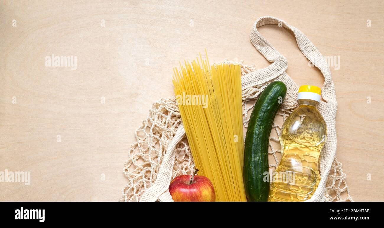 Lieferung von Lebensmitteln. Öko-Beutel Netz mit Nahrung: Öl, Spaghetti, Apfel, Gurke für Spenden während der Quarantäne. Essen auf dem Holzhintergrund Stockfoto