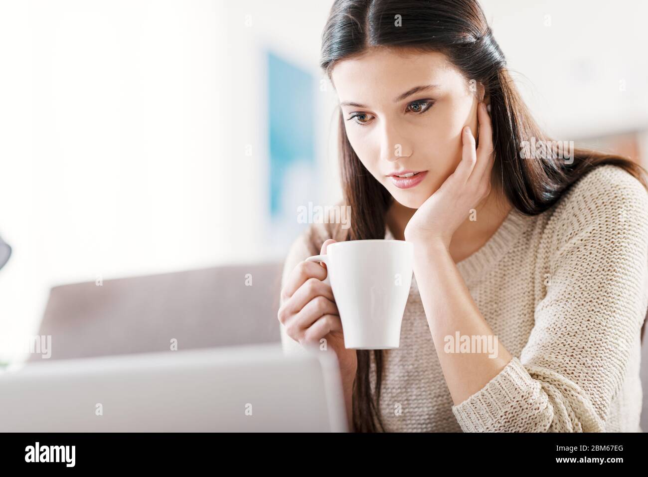 Junge Frau auf der Couch zu Hause sitzen und einen Film-streaming online auf ihrem Laptop, sie hält eine Tasse Kaffee Stockfoto