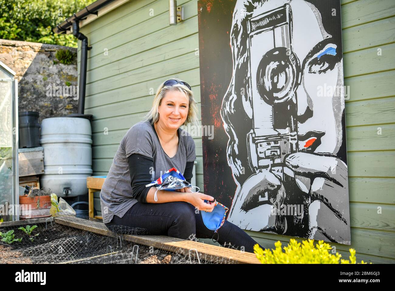 Die Künstlerin Shelley Dyer Gibbins, 47, aus dem Dorf Croscombe, in der Nähe von Wells, sitzt neben einem ihrer Werke mit dem Titel Cinegirl, Sie hat für alle 600 Einwohner des Dorfes Somerset Designer-Gesichtsmasken entworfen, sowohl aus medizinischem Maskenmaterial als auch aus einigen ihrer Kunstdruckmaterialien, die sie hinterlassen hatte. Stockfoto