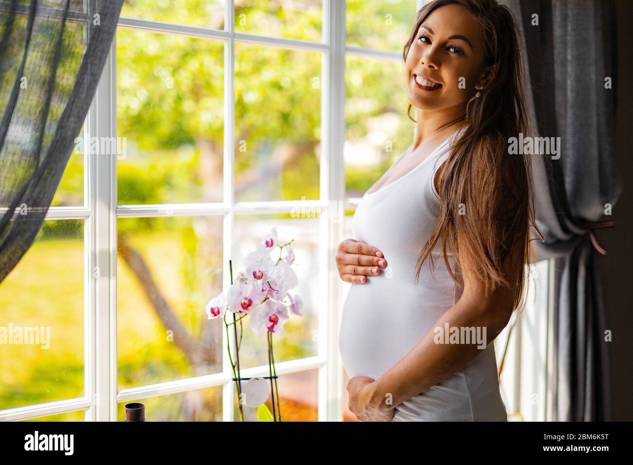 Lächelnde schwangere Frau, die am Fenster steht und ihren Bauch berührt Stockfoto