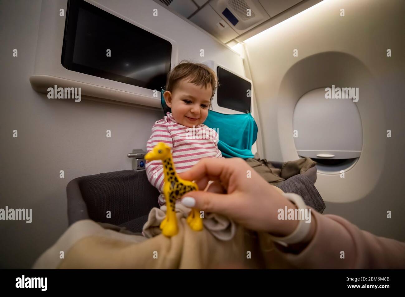 Glücklich lächelnd kleines süßes Kleinkind sitzt in der Babykassette eines Flugzeugs. Lacht und schaut auf die Hand ihrer Mutter mit einem Spielzeug in Unschärfe. Close-up, sof Stockfoto
