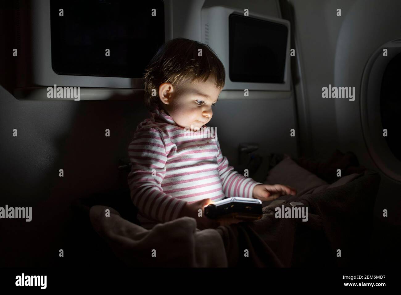 Kleines Kleinkind sitzt in speziellen Babygewanne auf einem Flugzeug in der Dunkelheit und spielt mit einer Taschenlampe. Stockfoto