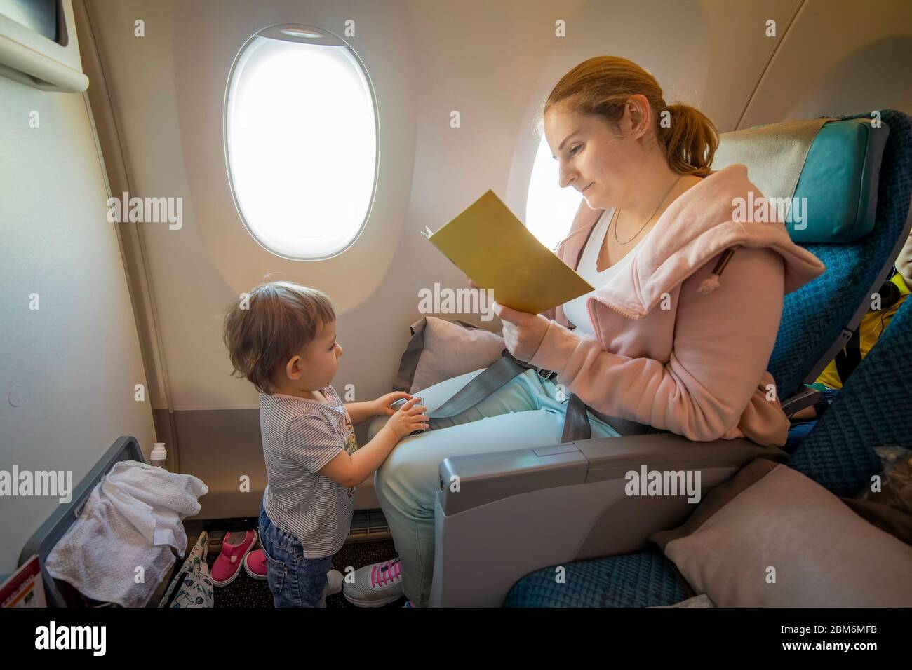 Junge schöne Mutter sitzt in einem Flugzeug Stuhl und liest ein Buch. Ihr kleines süßes Kleinkind steht vor ihr. Nahaufnahme, weiche Fokus, Draufsicht Stockfoto