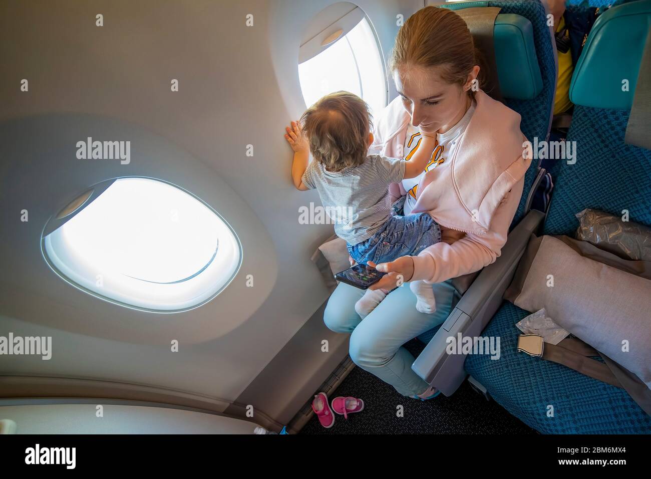 Junge schöne Mutter sitzt in einem Flugzeug Stuhl, ihr kleines süßes Baby sitzt in ihren Armen und schaut aus dem Fenster. Close-up, Soft-Focus, Stockfoto