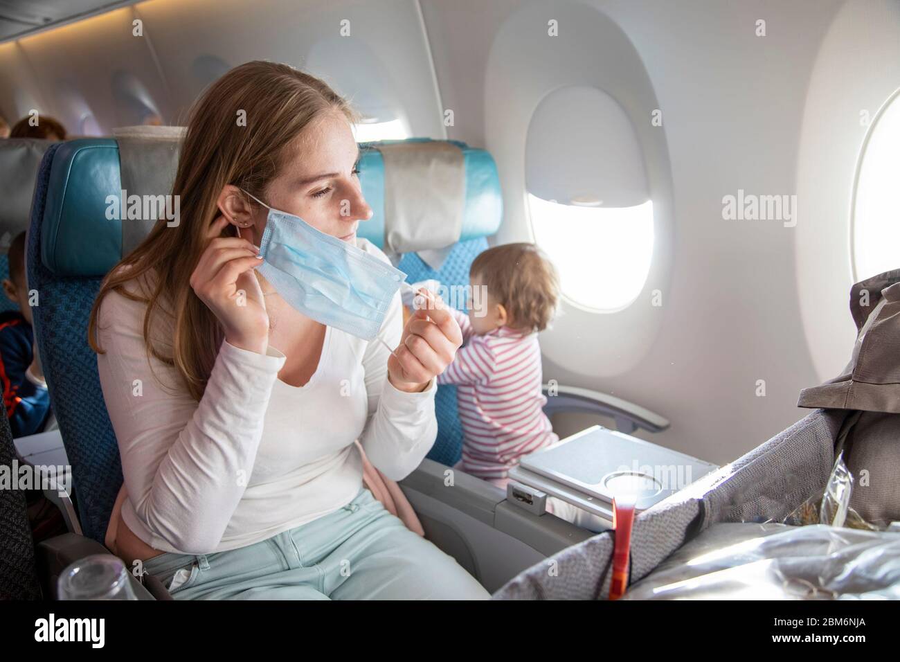 Konzeptreise mit einem Kind nach Asien, Angst vor Coronavirus covid-19. Junge schöne Mutter in einem Flugzeugsessel legt auf eine medizinische Atemmaske. Ein Schnitt Stockfoto