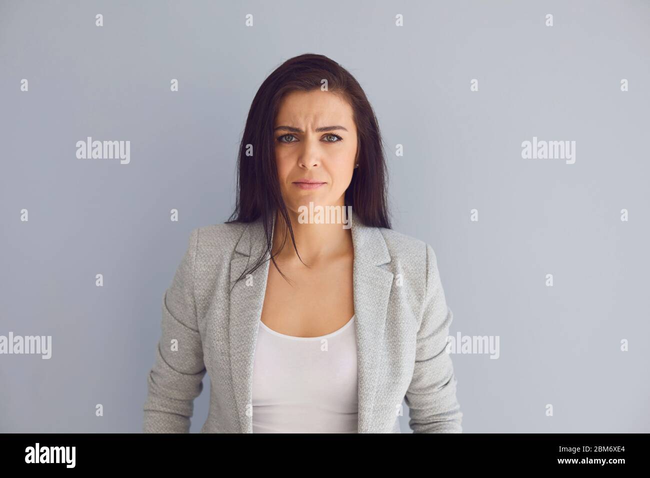 Junge beschäftigte Unternehmerin in stilvollem Outfit Blick auf Kamera und Stirnrunzeln mit ernsten Problemen, während vor grauem Hintergrund stehen Stockfoto