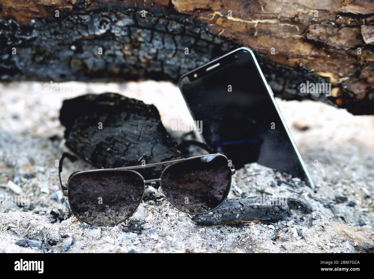 Telefon und Sonnenbrille in der Asche eines Lagerfeuers, verkohlte Holzstämme aus nächster Nähe Stockfoto
