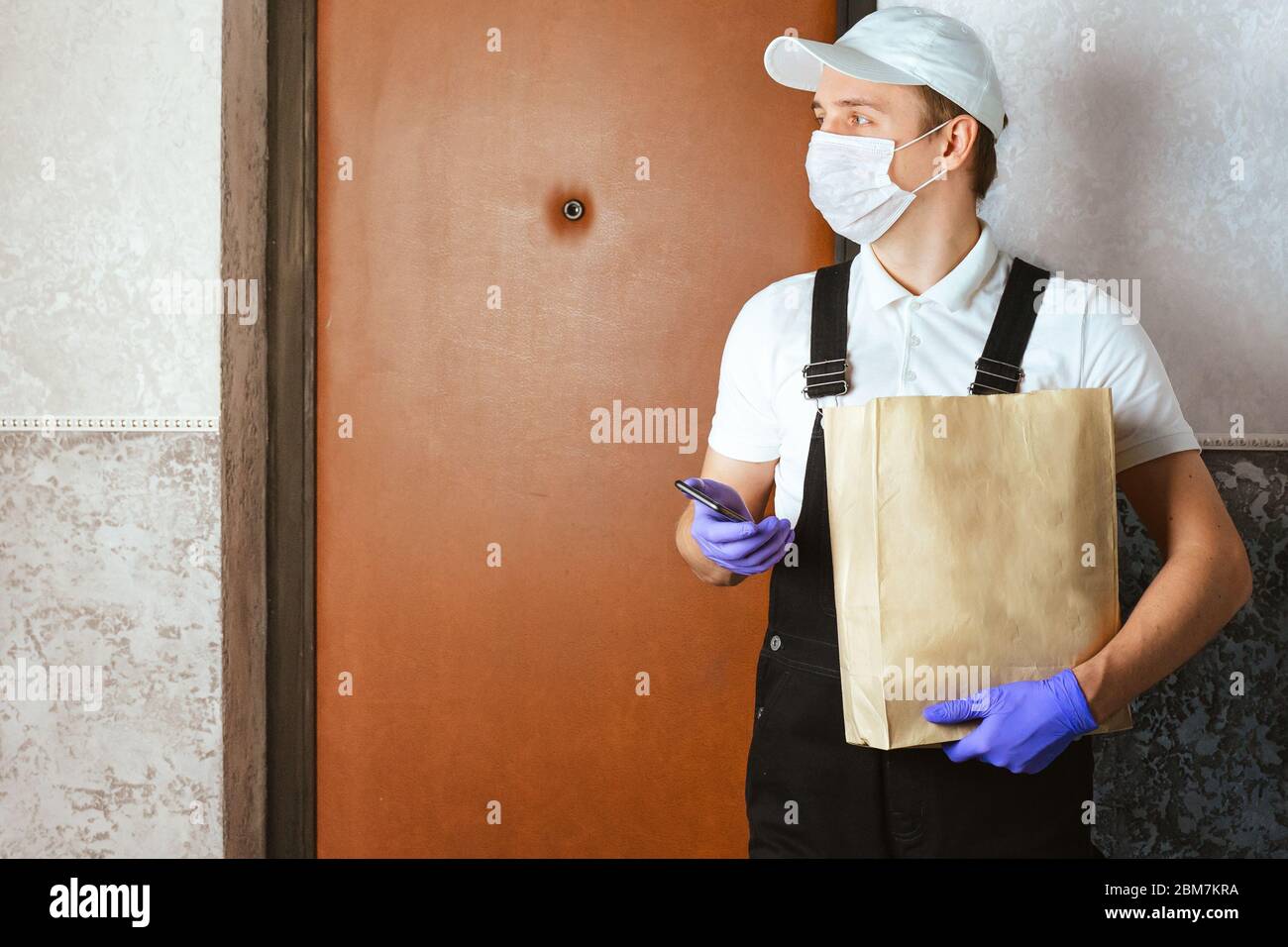 Männliche Kurier in chirurgischen Maske und medizinische Handschuhe mit Papiertüte mit Essen zum Mitnehmen. Volunteer Mann an der Tür liefern Online-Bestellung und überprüfen Telefon. Stockfoto