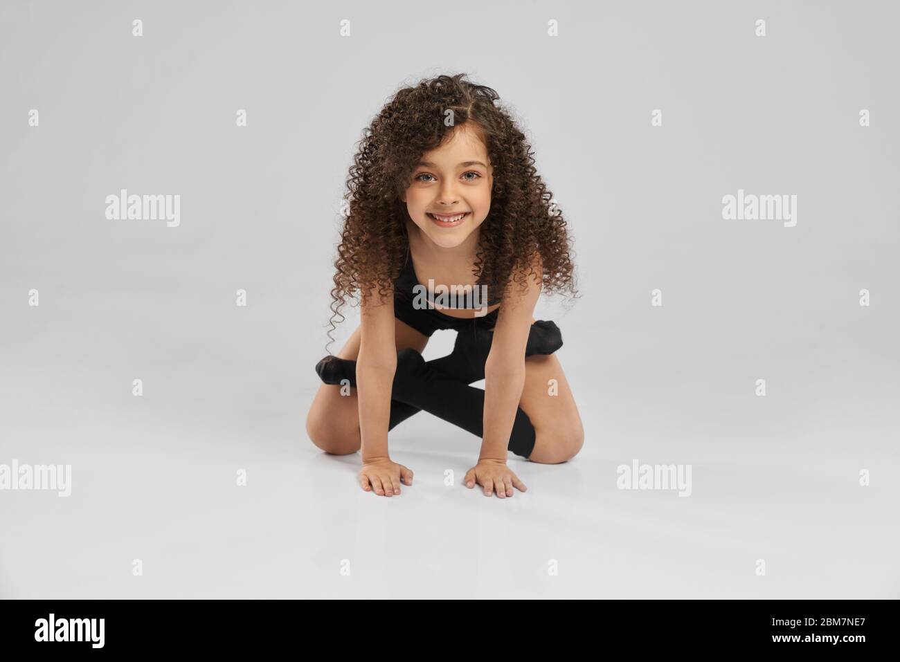 Nahaufnahme von attraktiven lächelndes Mädchen in schwarzer Sportkleidung und lange Socken stehen auf Knien in Lotus Pose des Bodens, isoliert auf grauem Studio Hintergrund. Kleine professionelle weibliche Turnerin mit lockigen Haaren. Stockfoto