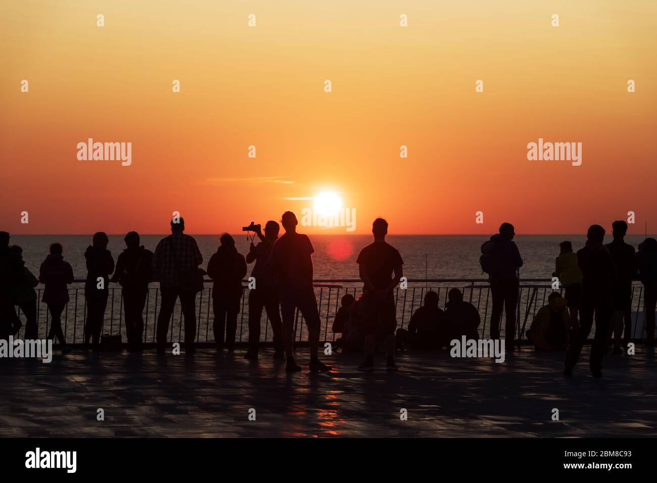 Silhouetten von Menschen gegen die untergehende Sonne auf dem Deck eines Kreuzfahrtschiffes. Ruhiges Meer und klarer orangener Himmel am Abend. Kreuzfahrt Urlaub Hintergrund Stockfoto