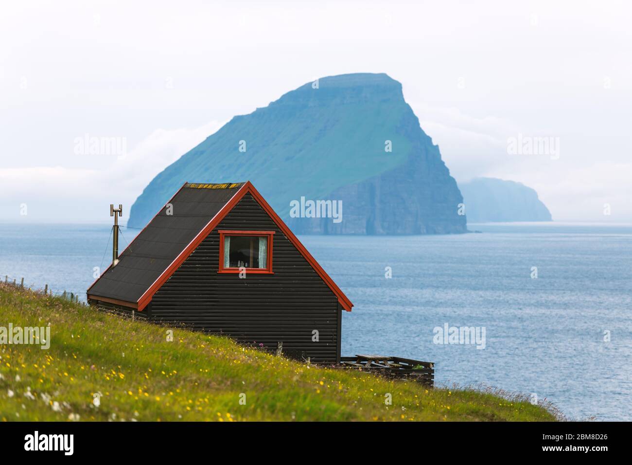 Schwarzes Haus auf dem berühmten färöischen Hexen Finger Trail und der Koltur Insel im Hintergrund. Dorf Sandavagur, Insel Vagar, Inseln der Färöer, Dänemark. Landschaftsfotografie Stockfoto