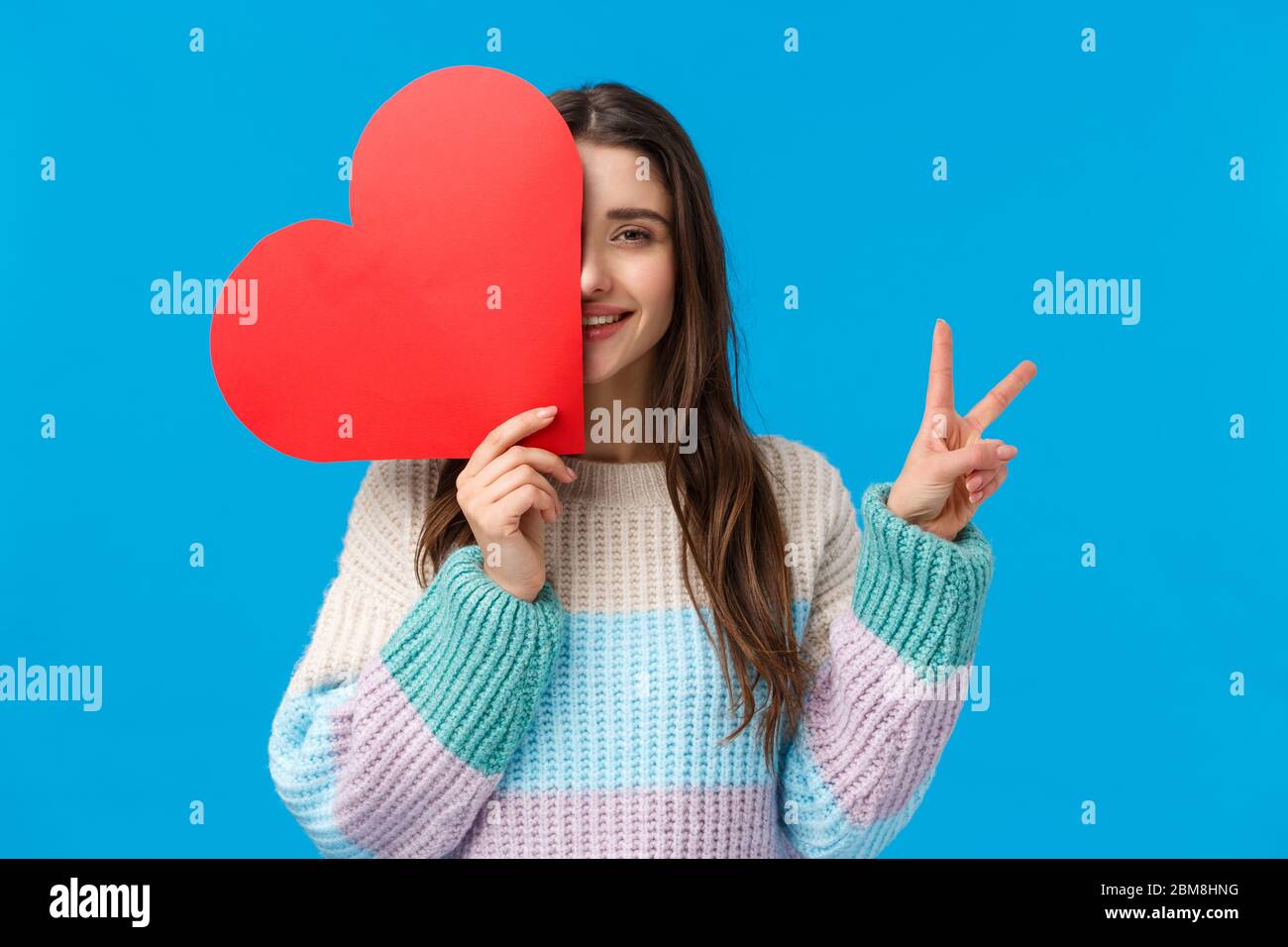 Taille-up-Porträt hübsche weibliche junge romantische Frau, große niedliche rote Herzkarte am Valentinstag, halten Sie sie über Gesicht, zeigen Sie ein Friedenszeichen und lächeln Stockfoto