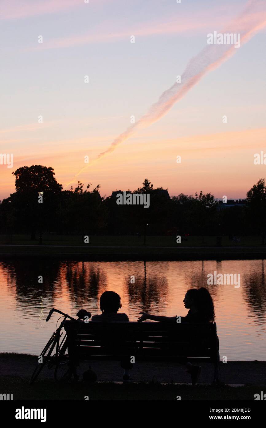 London, Großbritannien, 7. Mai 2020: Nach Tagen klaren Himmels verlassen immer mehr Flüge Dampfwege am Himmel. Auf Clapham Common spiegelte sich der dramatische Sonnenuntergang im Lond Pond wider und zwei Frauen saßen auf einer Bank und plauderten, vielleicht in Erwartung der versprochenen Lockdown-Regeln, die am Sonntag angekündigt werden. Anna Watson/Alamy Live News Stockfoto