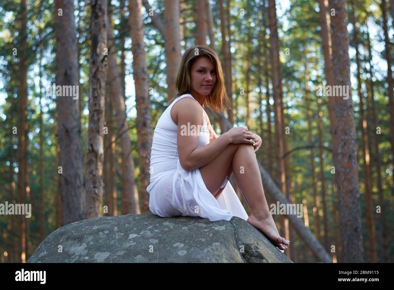 Ein Mädchen in einem weißen Kleid sitzt auf einem Felsen im Wald bei Sonnenuntergang Stockfoto