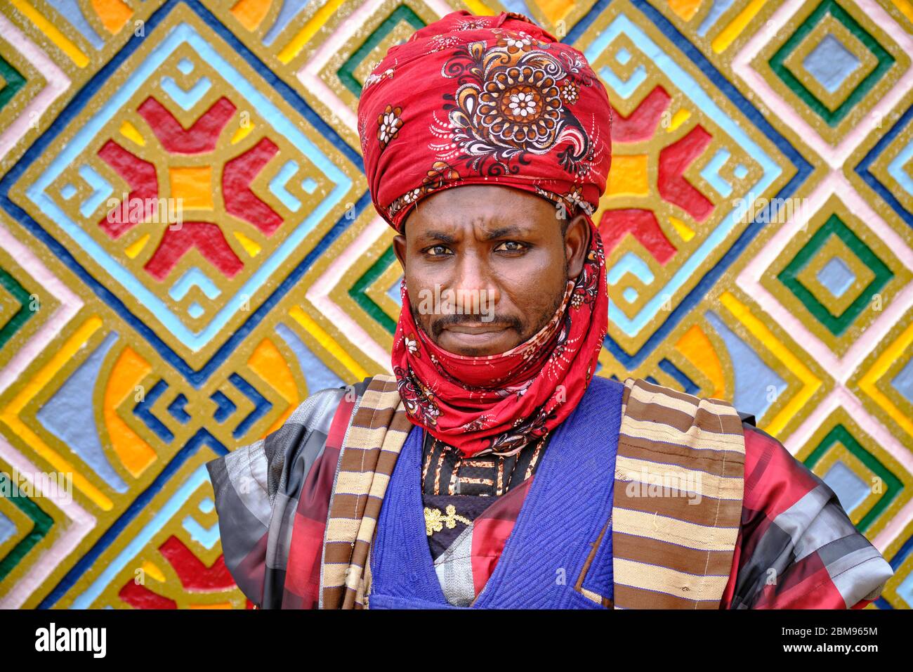 Diener des Emirpalastes in der Stadt Holländer in einer bunten traditionellen Uniform neben einer mit bunten Hausa-Motiven geschmückten Wand. Stockfoto