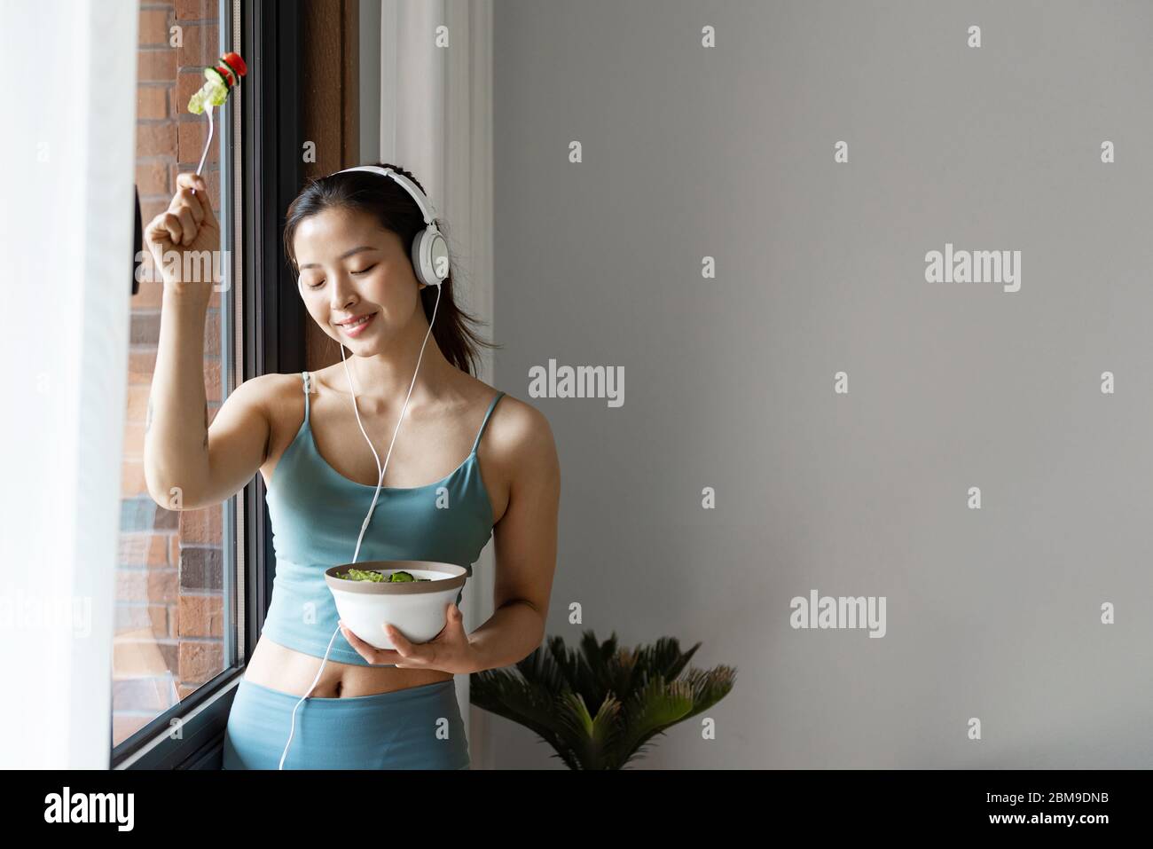 Eine junge asiatische Frau, die gesundes grünes Gemüse am Fenster isst Stockfoto