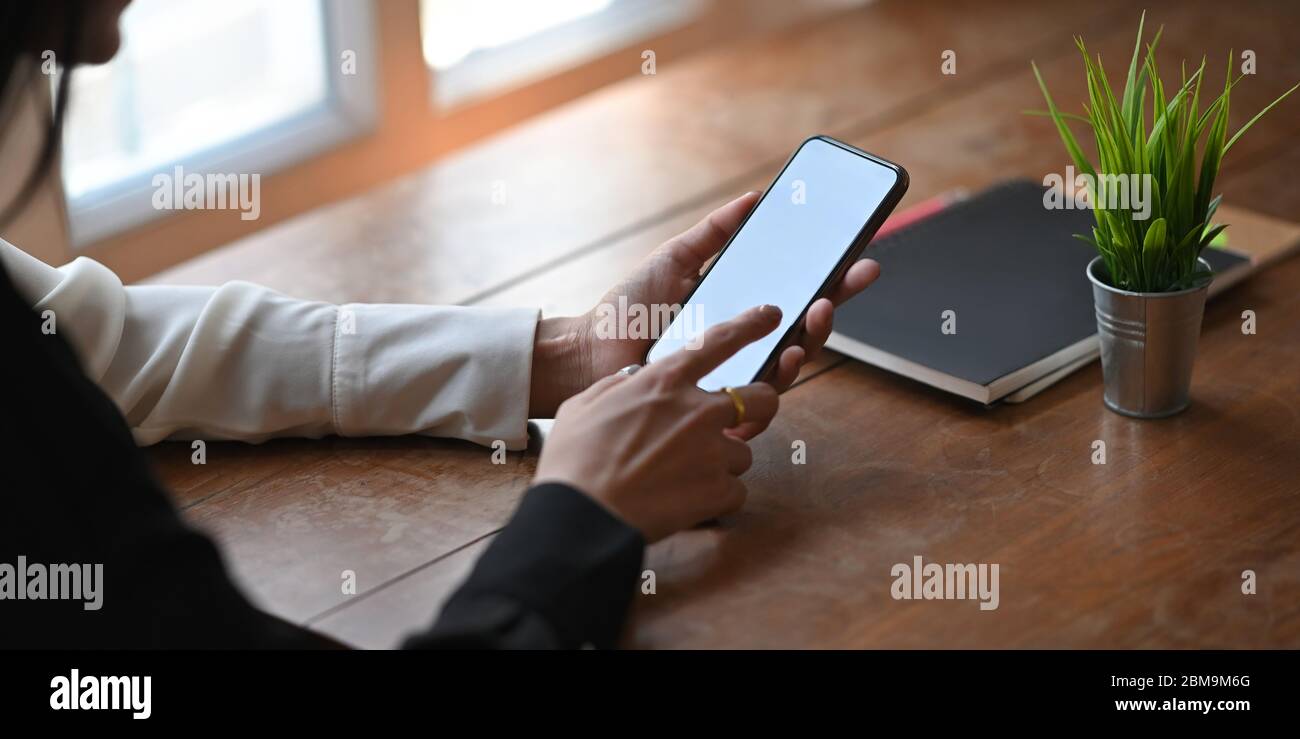 Zugeschnittenes Bild einer stilvollen Frau, die ein weißes Smartphone mit leerem Bildschirm hält und verwendet, während sie am Holzschreibtisch über dem komfortablen Wohnzimmer sitzt Stockfoto