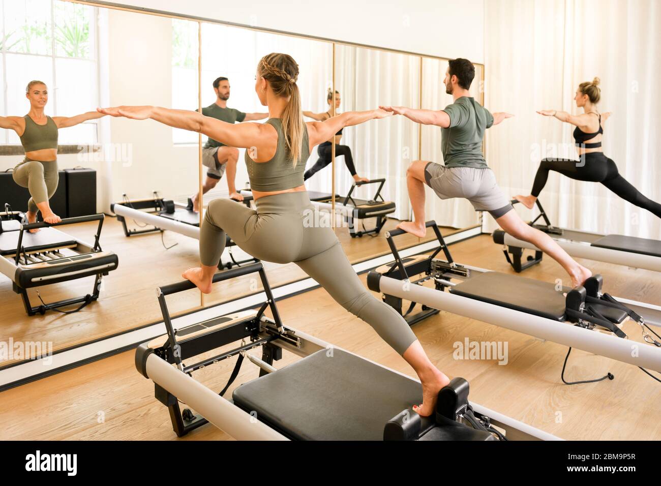 Pilates-Klasse von Athleten, die eine stehende Ausfallschritt Übung auf Reformerbetten in einem Wandspiegel in einem High-Key-Fitnessstudio in einem Gesundheits-und Fitness-concep reflektiert Stockfoto