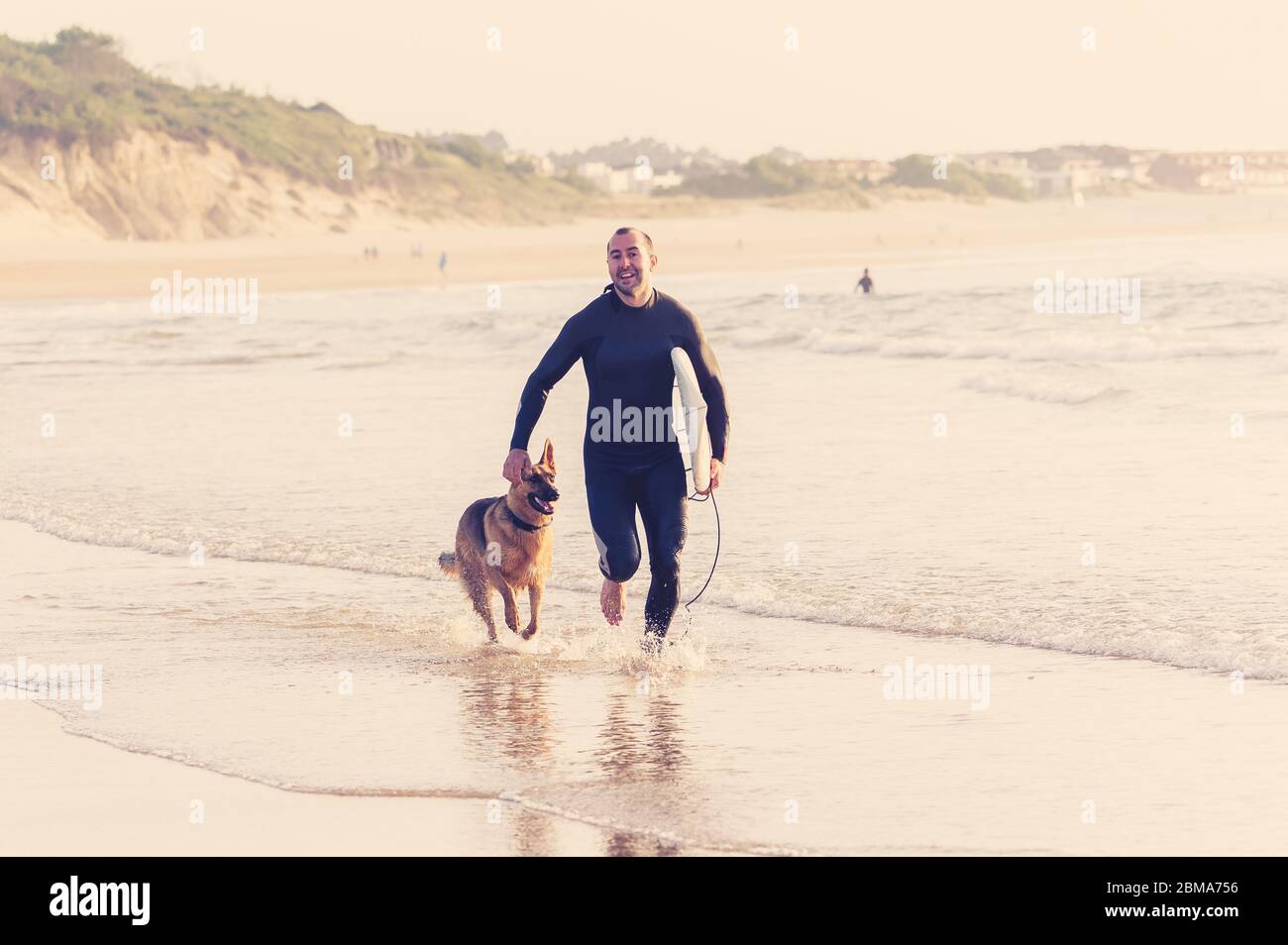 Surfer Spaß mit besten Freund deutschen Schäferhund laufen und spielen auf hundefreundlichen Strand bei Sonnenuntergang. Sommer Spaß Surfen Urlaub mit Ihrem Hund, Haustier Stockfoto