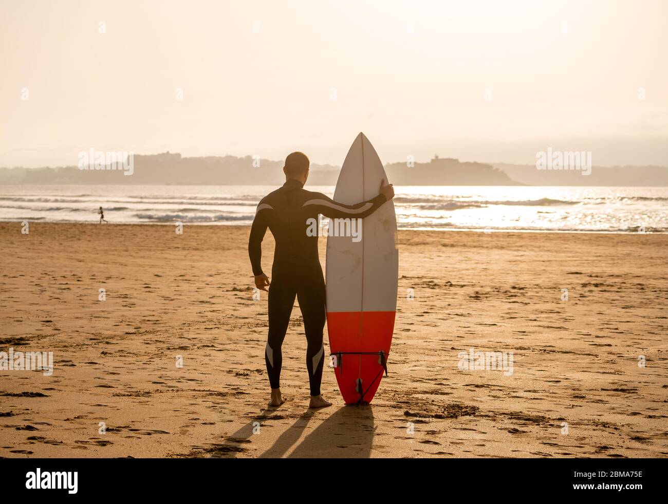 Rückansicht des Surfers mit Surfbrett am Strand bei Sonnenuntergang oder Sonnenaufgang. Silhouette des Surfmanns, der auf das Meer schaut und auf hohe Wellen wartet. Extrem Stockfoto