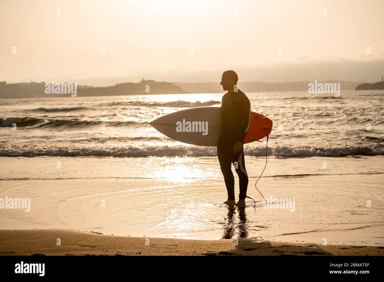 Starker Surfer mit Surfbrett am Strand beim Sonnenauf- oder -Untergang. Silhouette des Surfmanns stehend auf die Wellen des Ozeans. Im Freien, extremer Spor Stockfoto