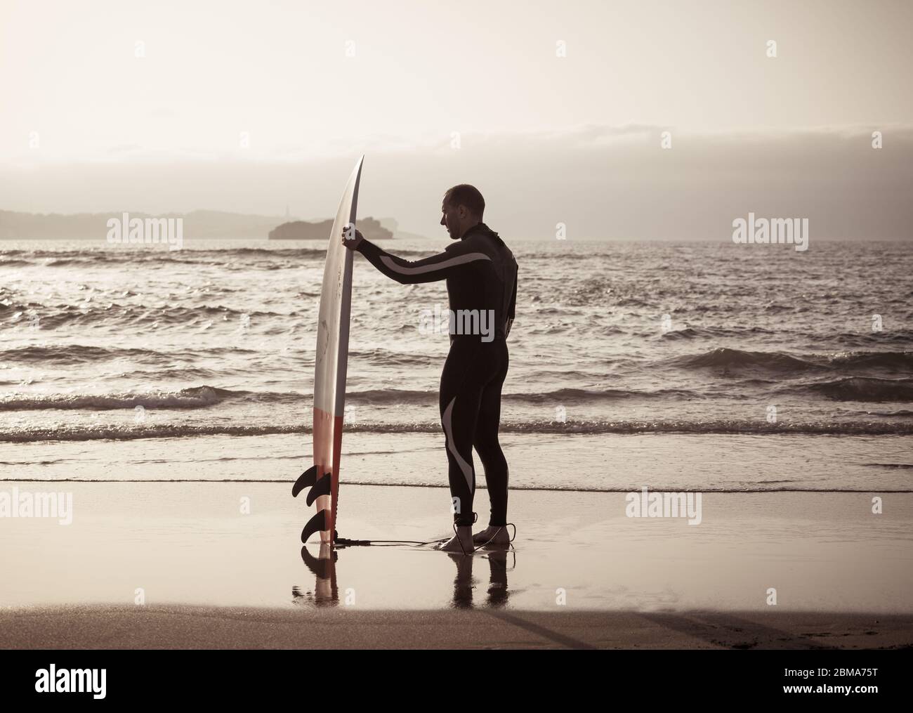 Rückansicht des Surfers mit Surfbrett am Strand bei Sonnenuntergang oder Sonnenaufgang. Silhouette des Surfmanns, der auf das Meer schaut und auf hohe Wellen wartet. Extrem Stockfoto