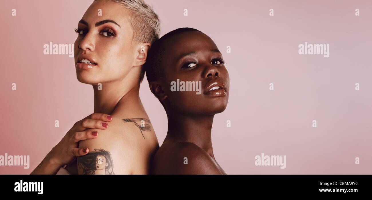 Selbstbewusste verschiedene weibliche Modelle stehen zurück an Rücken und Blick auf die Kamera. Zwei multiethnische Frauen stehen zusammen auf beigefarbenem Hintergrund. Stockfoto