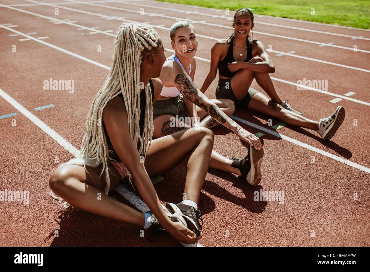 Glückliche Gruppe von weiblichen Athleten auf einer Laufstrecke sitzen und Stretching Beine. Frauen Läufer machen Aufwärmübungen im Stadion. Stockfoto