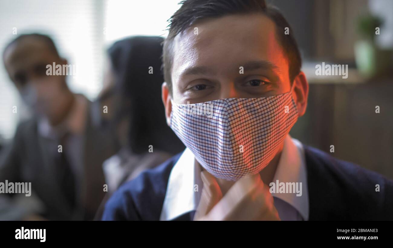 Positiver Geschäftsmann korrigiert Schutzmaske auf seinem Gesicht. Porträt des jungen Mannes im Büro von Neonlicht vor dem Hintergrund der Menschen beleuchtet Stockfoto