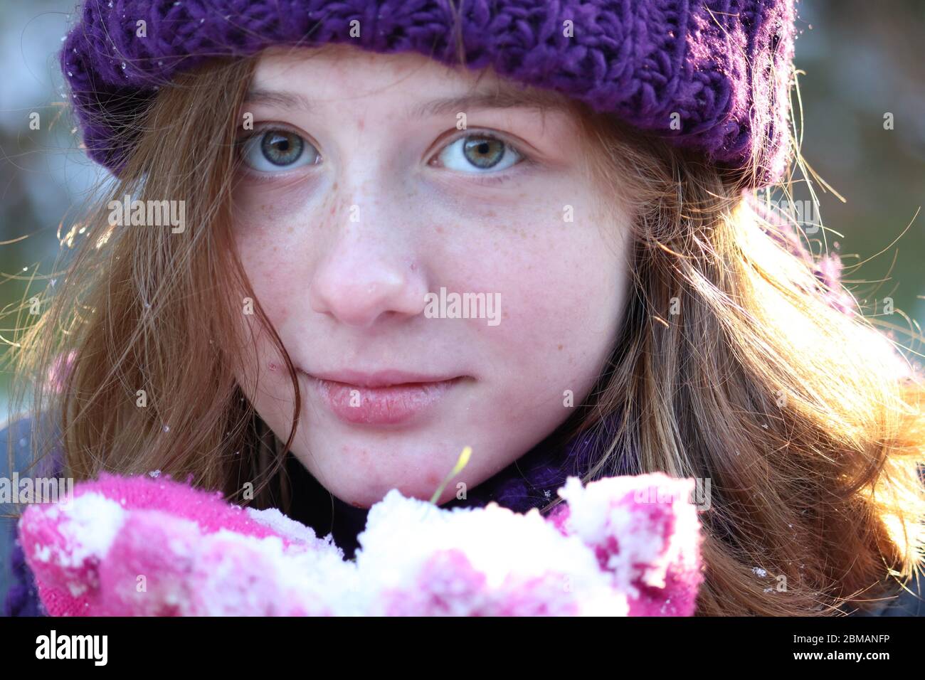 Junge, blauäugige Mädchen mit violettem Strickmütze und rosa Handschuhen, die im Winter Schnee halten und Sonne auf ihrem Haar in Lancaster County, Pennsylvania, scheint Stockfoto