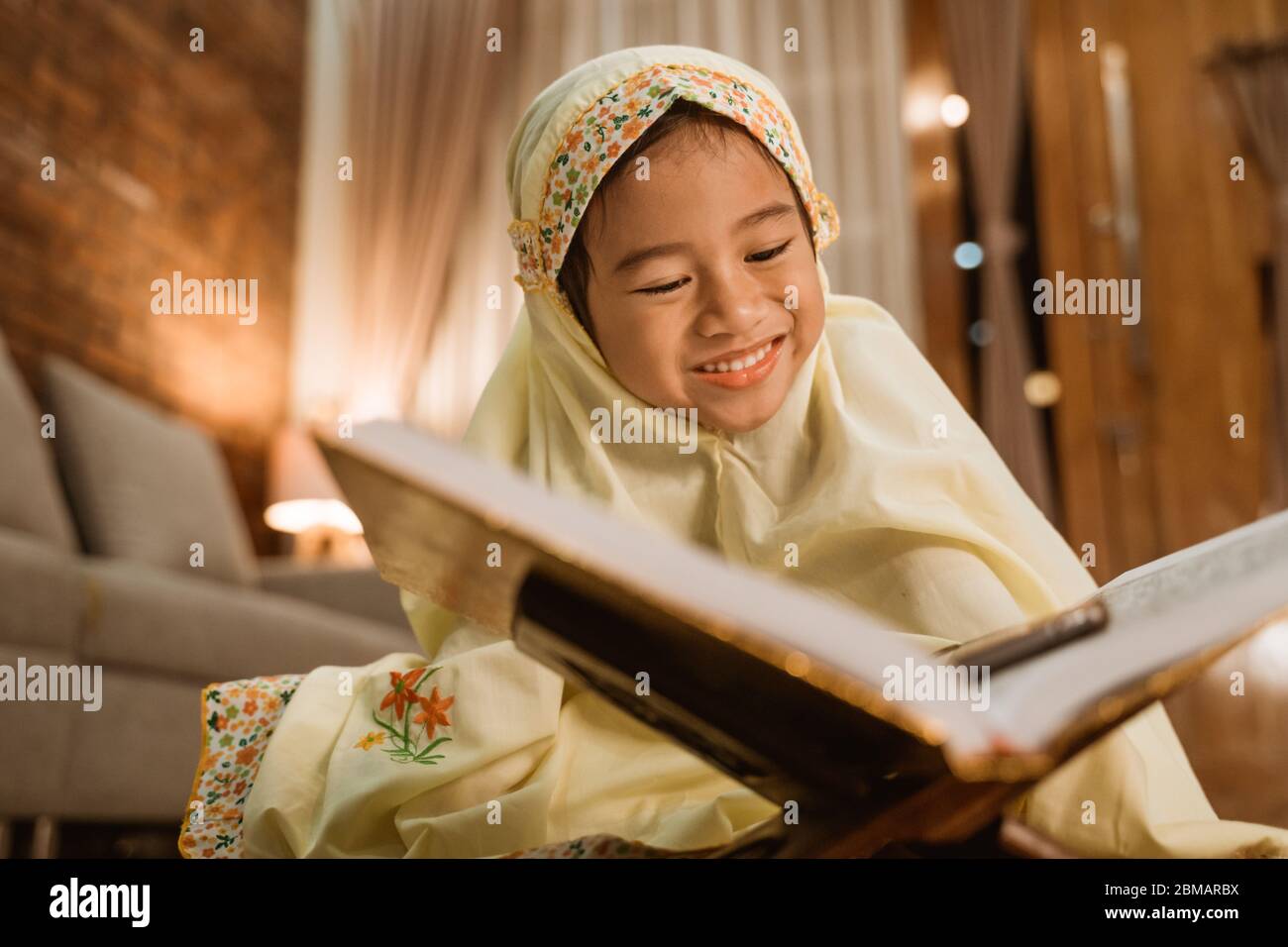 Schönes kleines Mädchen, das den koran liest und allein zu Hause einen muslimischen Hijab trägt Stockfoto