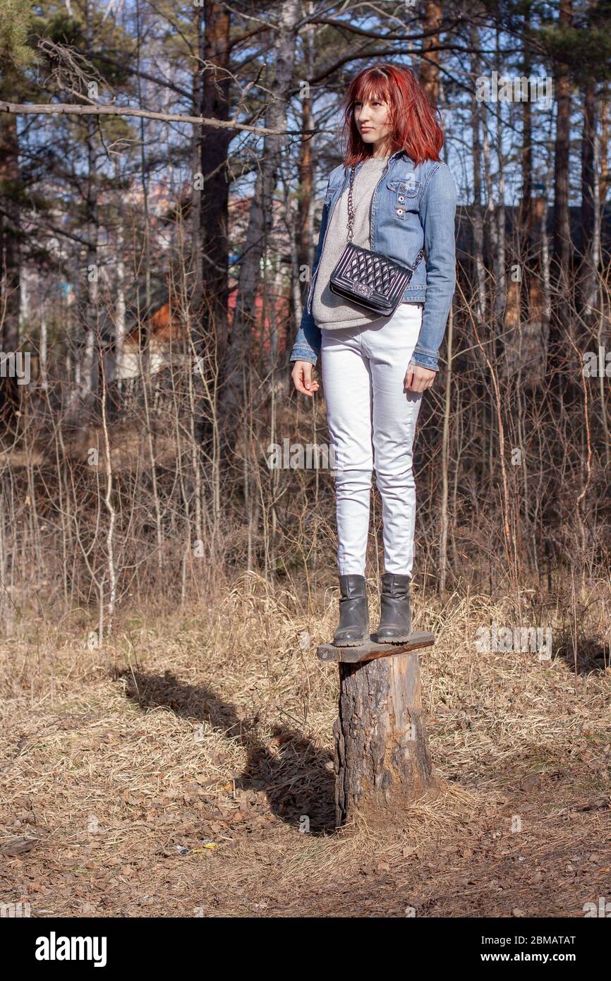 Ein junges rothaariges Mädchen steht auf einem Stumpf im Wald. Europäisches  Erscheinungsbild. Jeansjacke und weiße Hose. Handtasche auf der Schulter.  Vertikal Stockfotografie - Alamy