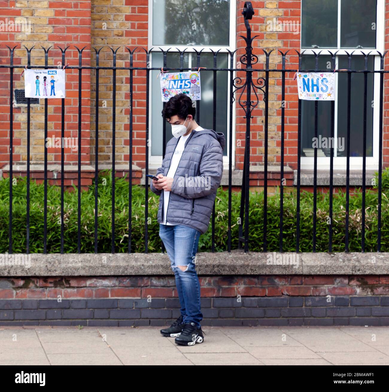 Der junge Mann, der eine Gesichtsmaske trug, benutzte sein Mobiltelefon außerhalb des Lewisham Krankenhauses während der COVID-19 Pandemie. 'Danke NHS'-Schilder hängen hinter ihm am Geländer Stockfoto