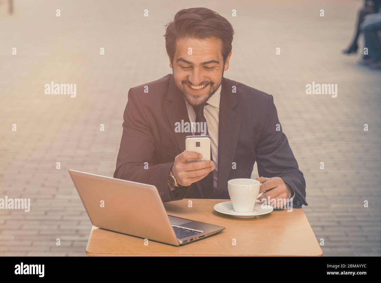 Geschäftsmann nutzt Smartphone arbeiten im Freien. Glücklicher Mann schaut auf das Handy sitzen am Tisch mit Computer und Tasse Kaffee auf ihm auf der Straße Café Stockfoto