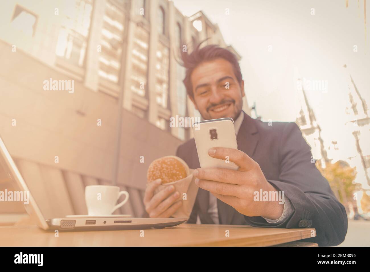 Geschäftsmann isst Hamburger mit Telefon auf der Straße. Lächelnder Mann im Anzug, der in der Fast-Food-Cafeteria auf der Straße einer modernen europäischen Stadt isst. Getönte Stockfoto