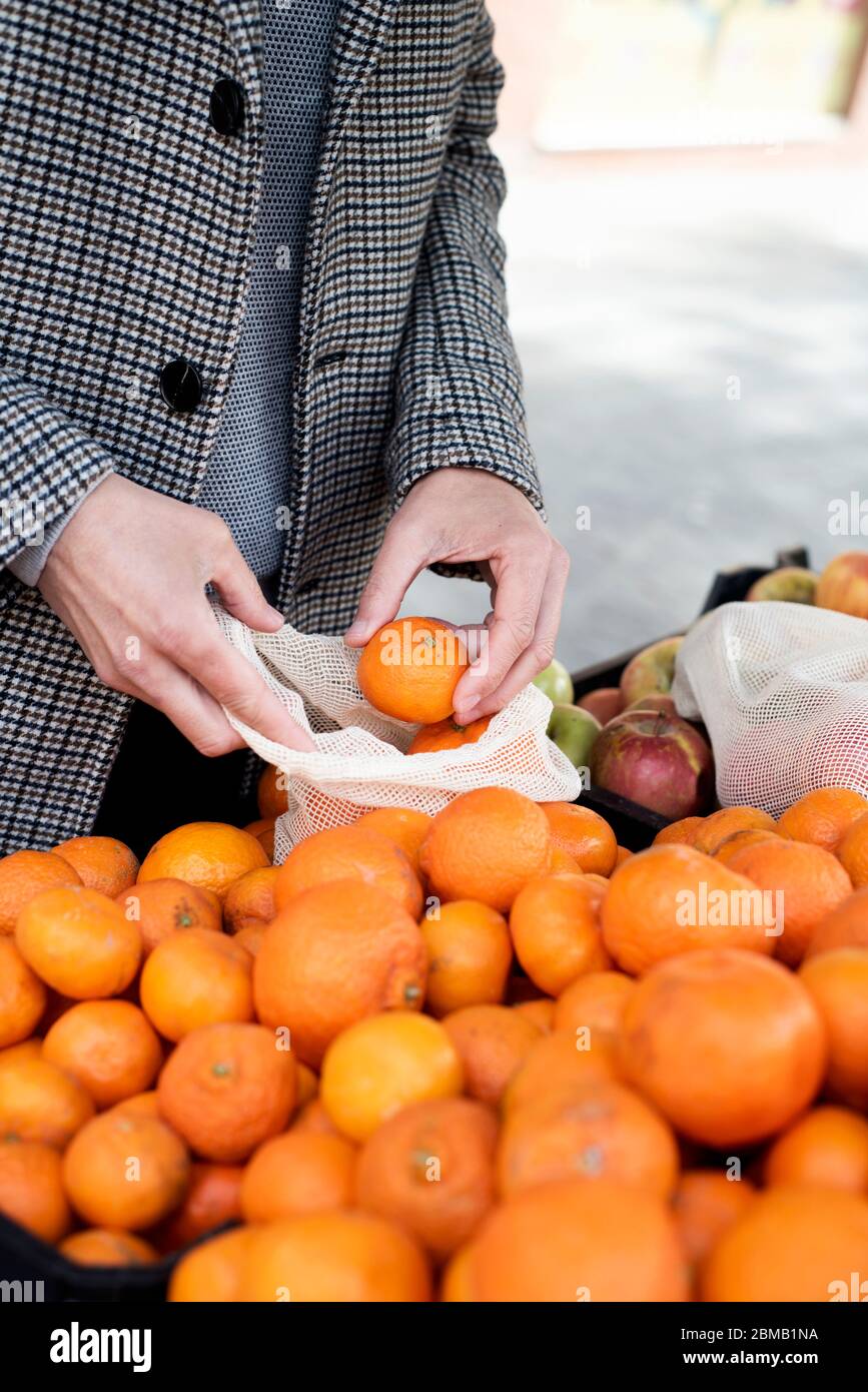 Nahaufnahme eines Mannes, der in einem Greengrocker einkauft und einige Mandarinorangen in einen wiederverwendbaren Textil-Netzbeutel einbringt, um die Kunststoffverschmutzung zu verringern Stockfoto
