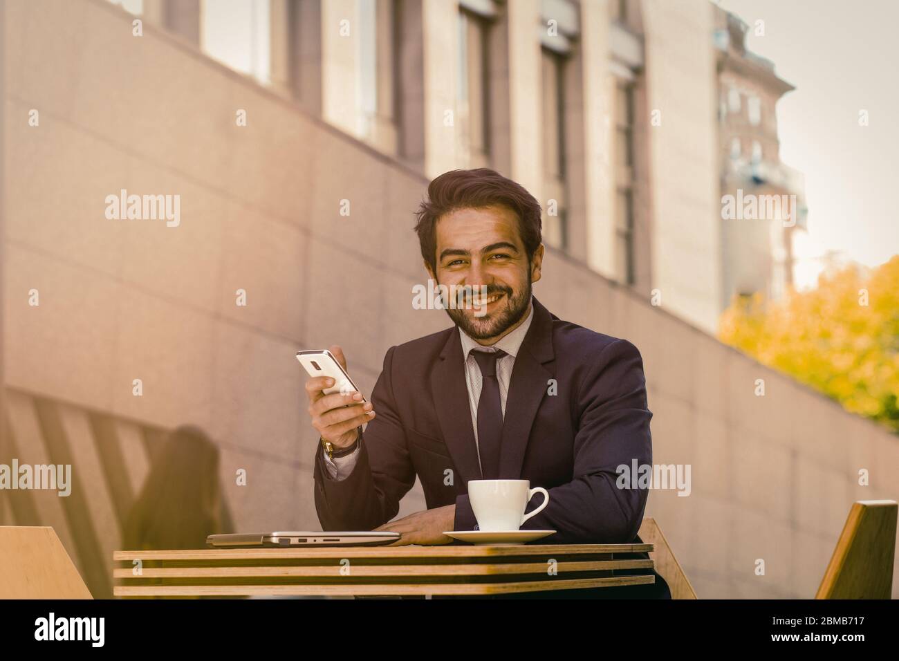 Geschäftsmann, der im Freien ein Mobiltelefon verwendet. Erfolgreicher Mann toothy lächelt Smartphone während am Tisch sitzen mit einer Tasse Kaffee auf ihm in der Stadt Stockfoto