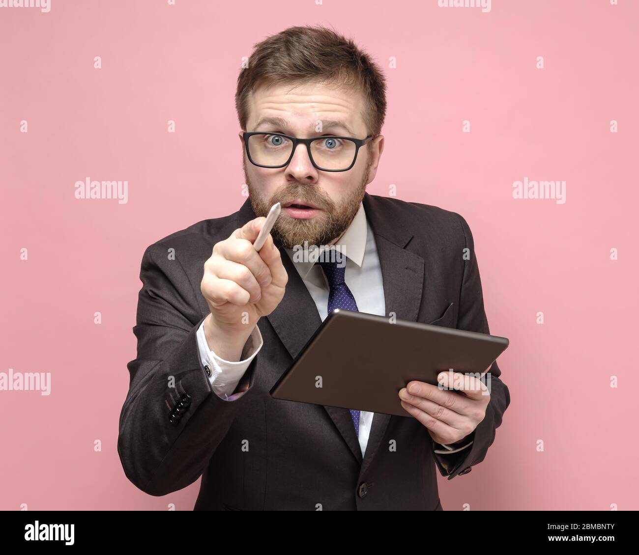 Kaukasischer Geschäftsmann in einem Anzug mit Tablet sieht neugierig und zeigt mit einem Stift auf die Kamera. Stockfoto