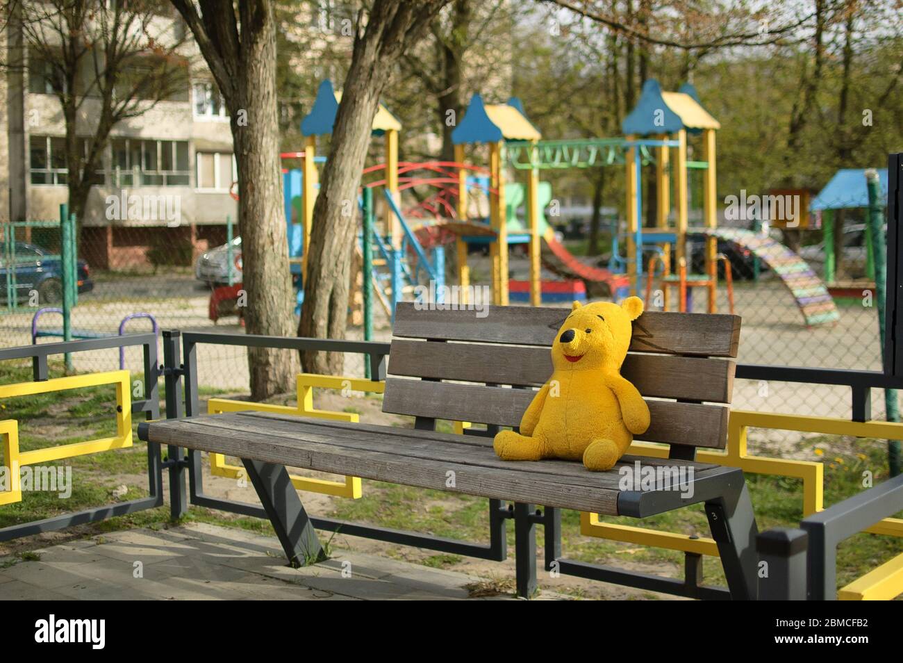 2020.04.18 Lviv, Ukraine. Gelbbär Spielzeug sitzt auf der Bank in der Nähe von leeren geschlossenen Spielplatz im Freien ohne Kinder wegen Quarantäne Maßnahmen Stockfoto